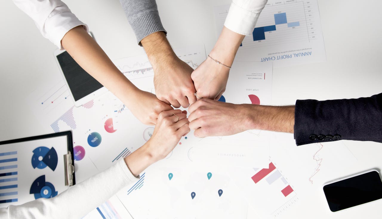 hero-img-02 Colleagues celebrate success with a fist bump over financial charts depicting teamwork and unity.