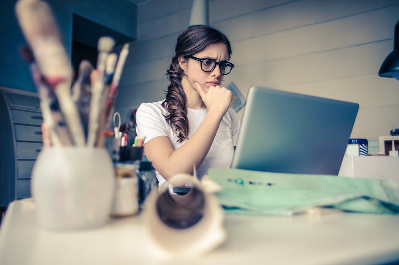 bts-img-02 Young woman with glasses deeply focused on a laptop surrounded by art supplies in a home office.