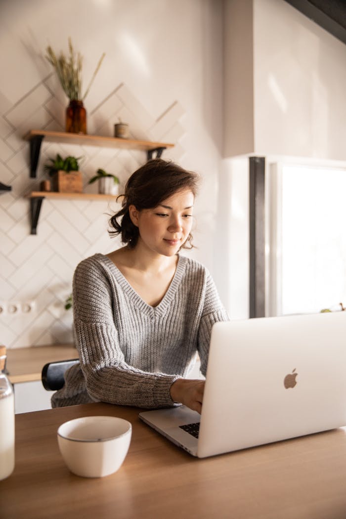 my-work-03 Young female in gray sweater sitting at wooden desk with laptop and bottle of milk near white bowl while browsing internet on laptop during free time at home